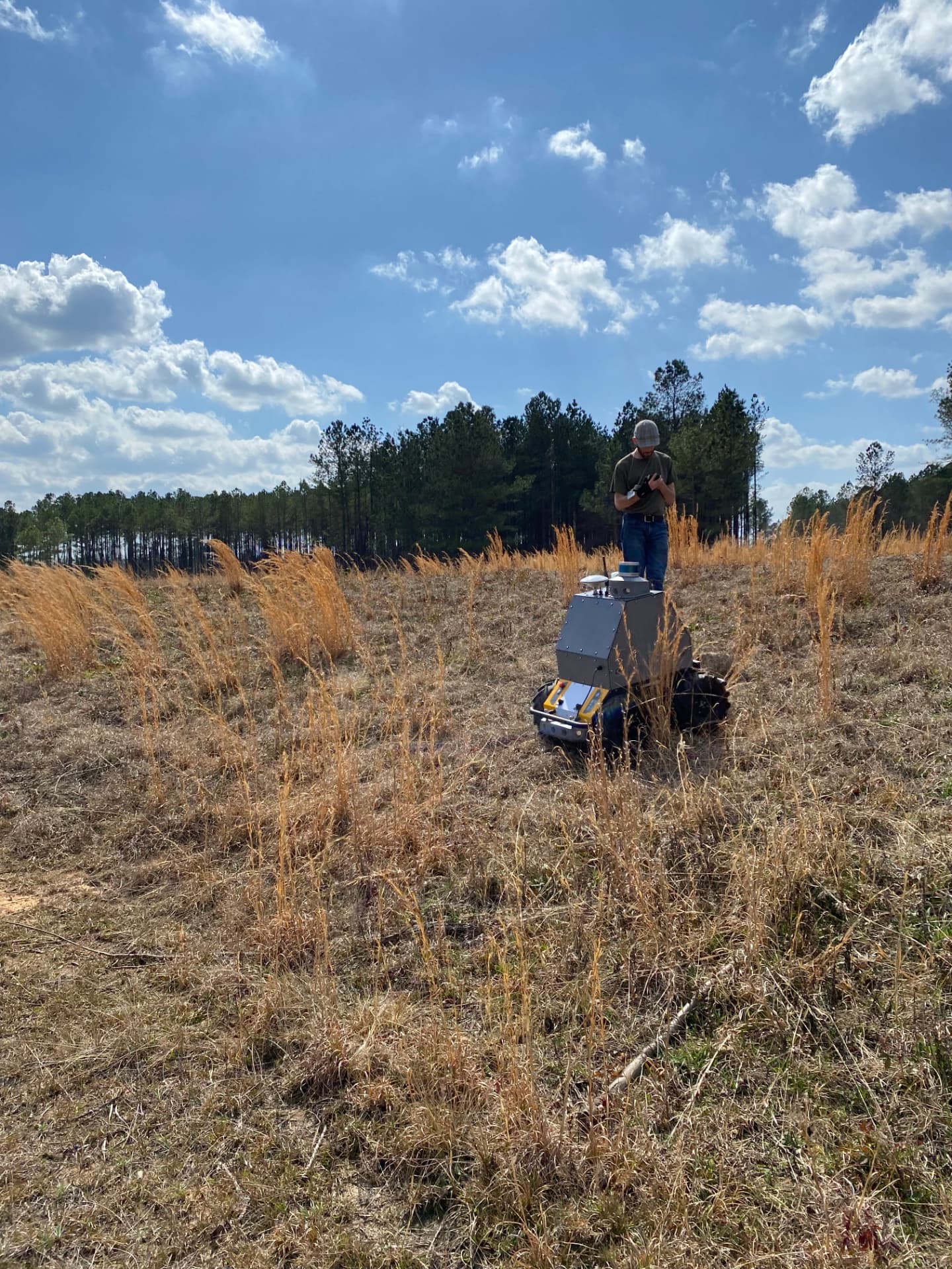 Remote-operated tracked equipment cutting overgrown vegetation across a field while an operator monitors it
