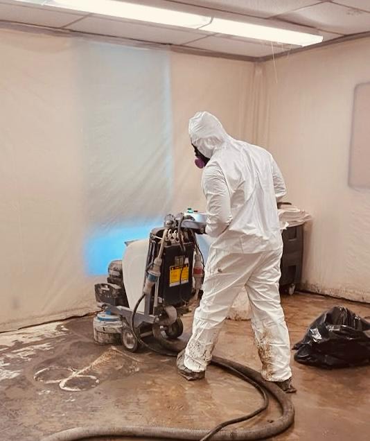 Worker in a full white protective suit and respirator operating a floor removal machine inside a plastic-sheeted containment area