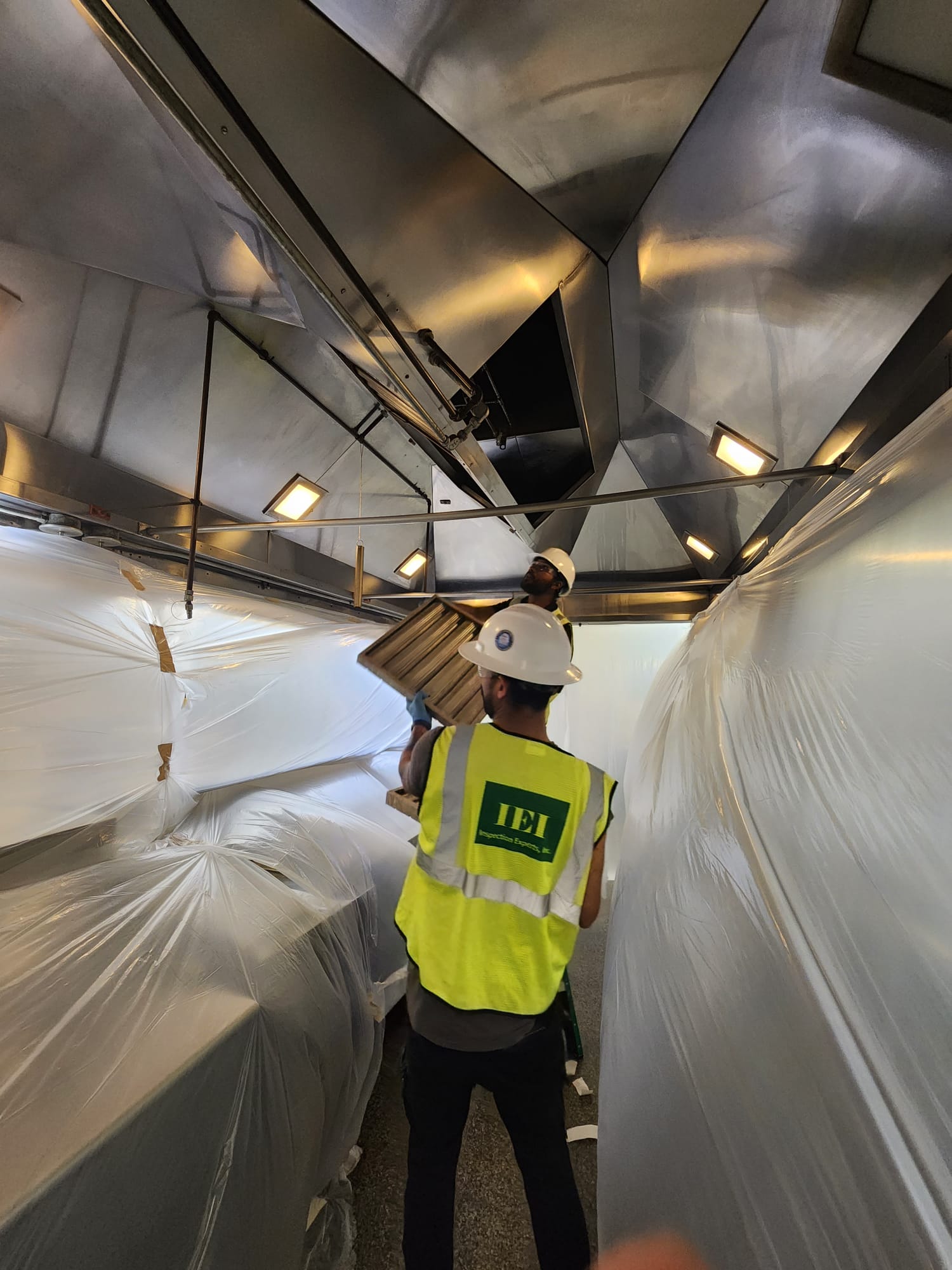 IEI crew in hard hats and IEI vests working under containment sheeting inside a commercial kitchen exhaust hood
